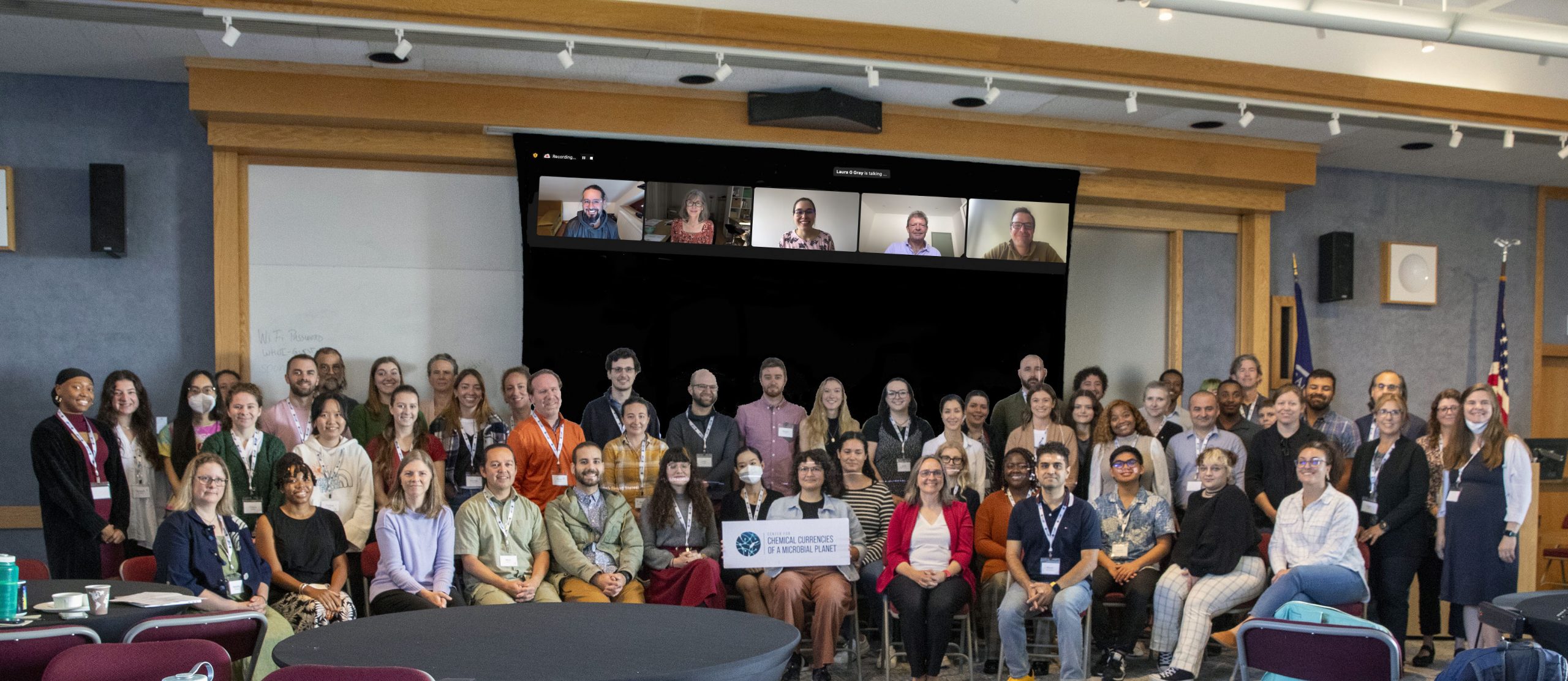 A group of in-person CoMP members gather in front of a projector screen displaying remote participants who joined the meeting virtually. 