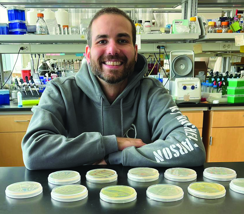 Dr. Frank Xavier Ferrer Gonzalez stands behind a lab bench holding petri dishes containing cultures of bacteria.
