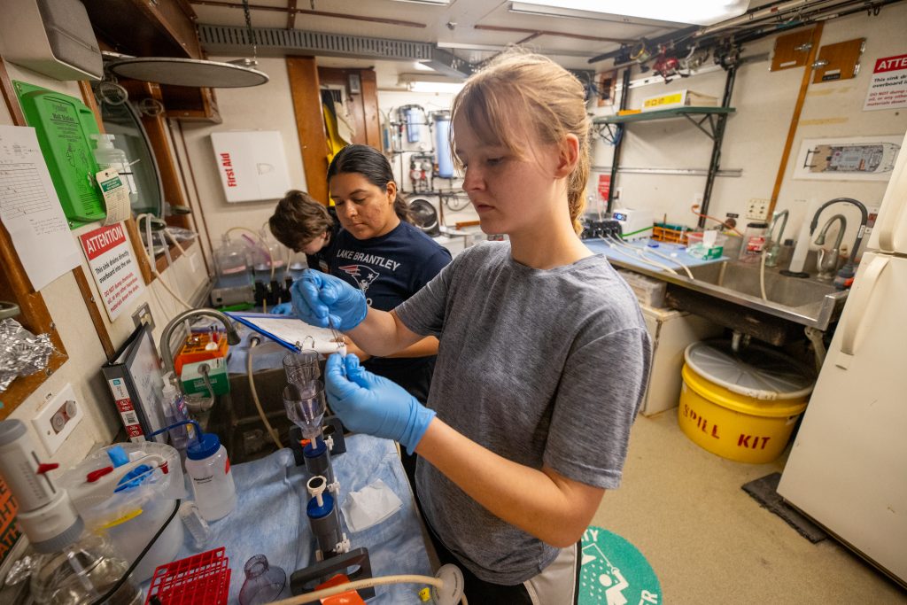 Two researchers stand in front of a bench within a ship-board laboratory in order to filter seawater.