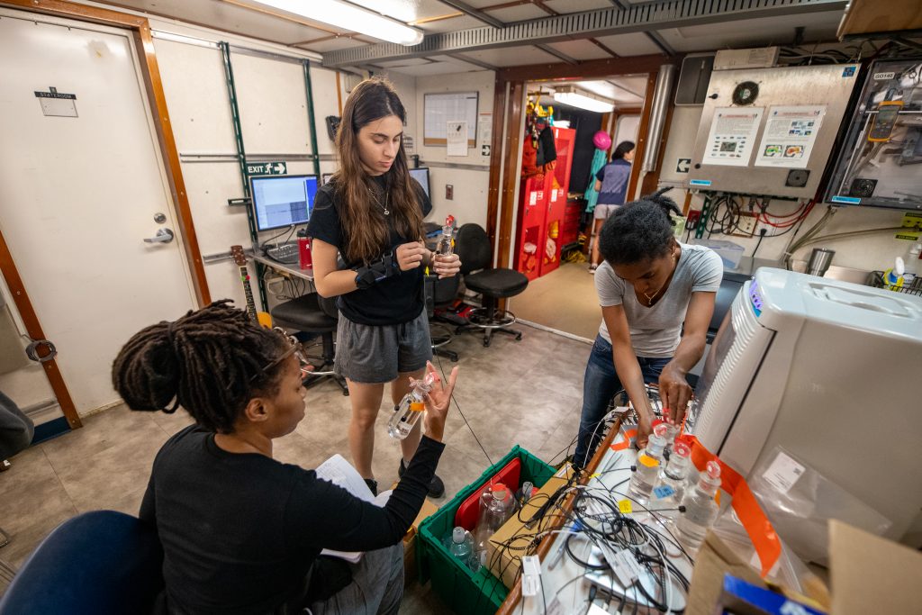 Three researchers are located next to a table covered in science equipment in a ship-board lab. One researcher is sitting, and two are standing. Two researchers are holding glass bottles.