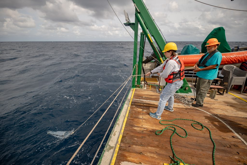 A researcher lowers a rope over the side of the vessel while standing on the deck of a ship next to a marine technician.