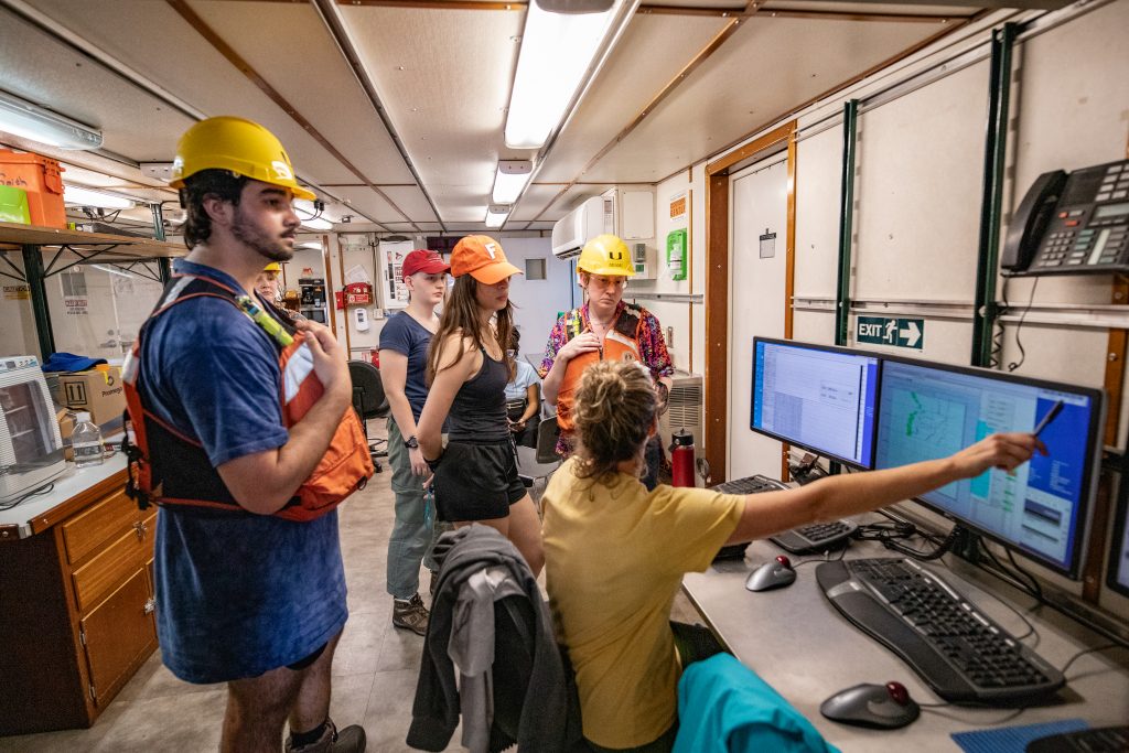 A group of researchers stand in front of two computer monitors in a ship-board. One researcher, sitting in front of the computer monitors, points to the screen.