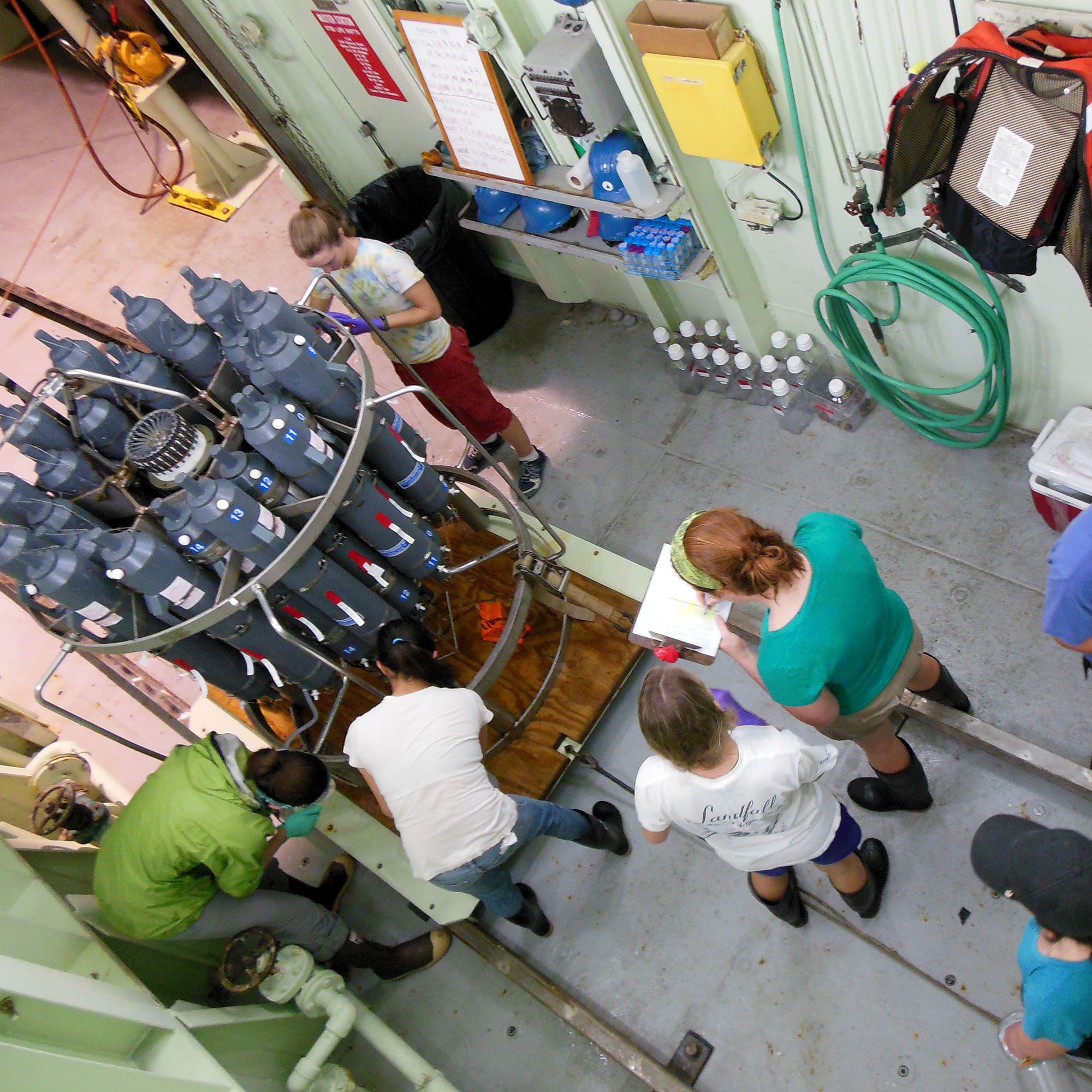 Five researchers are surrounding a niskin ctd rosette after samples have been collected on a research vessel. Three people are collected seawater directly from the niskin bottles while two people are looking at a datasheet.