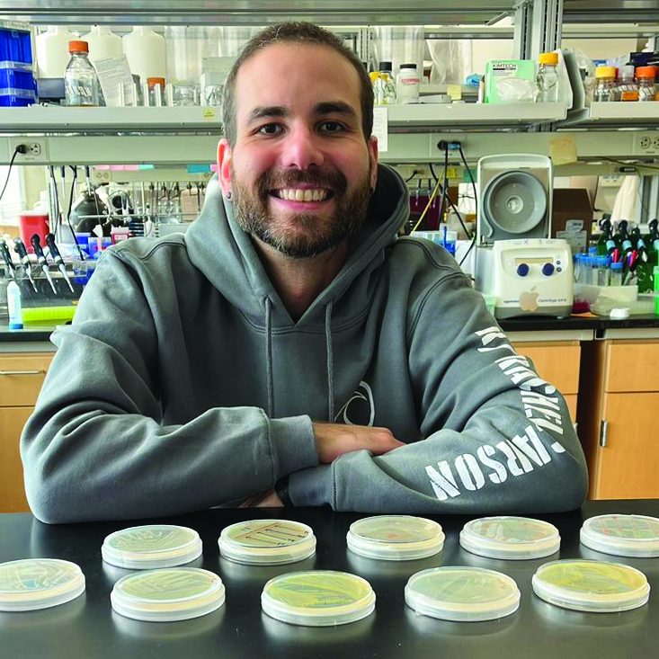 Dr. Frank Xavier Ferrer Gonzalez stands behind a lab bench holding petri dishes containing cultures of bacteria.